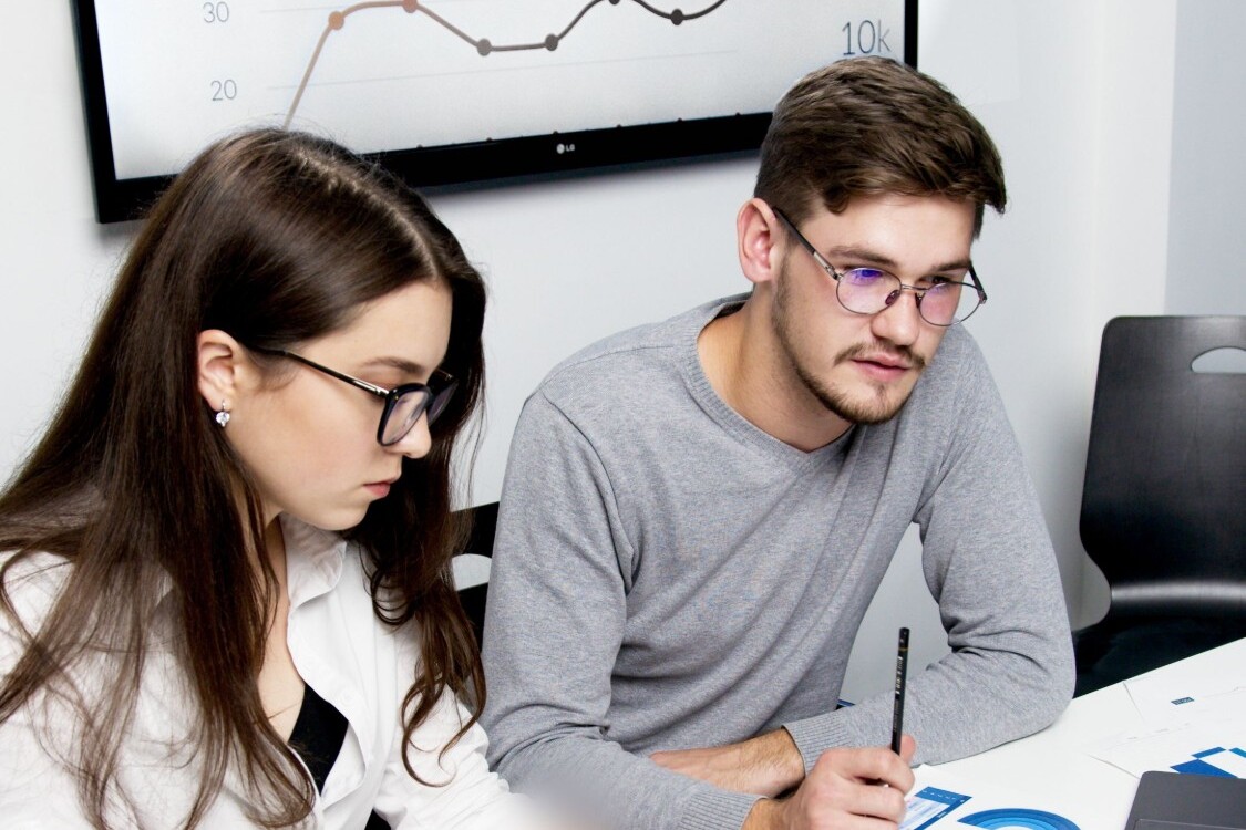 Dois alunos a analisar gráficos e dados num ecrã, com apontamentos e documentos na mesa