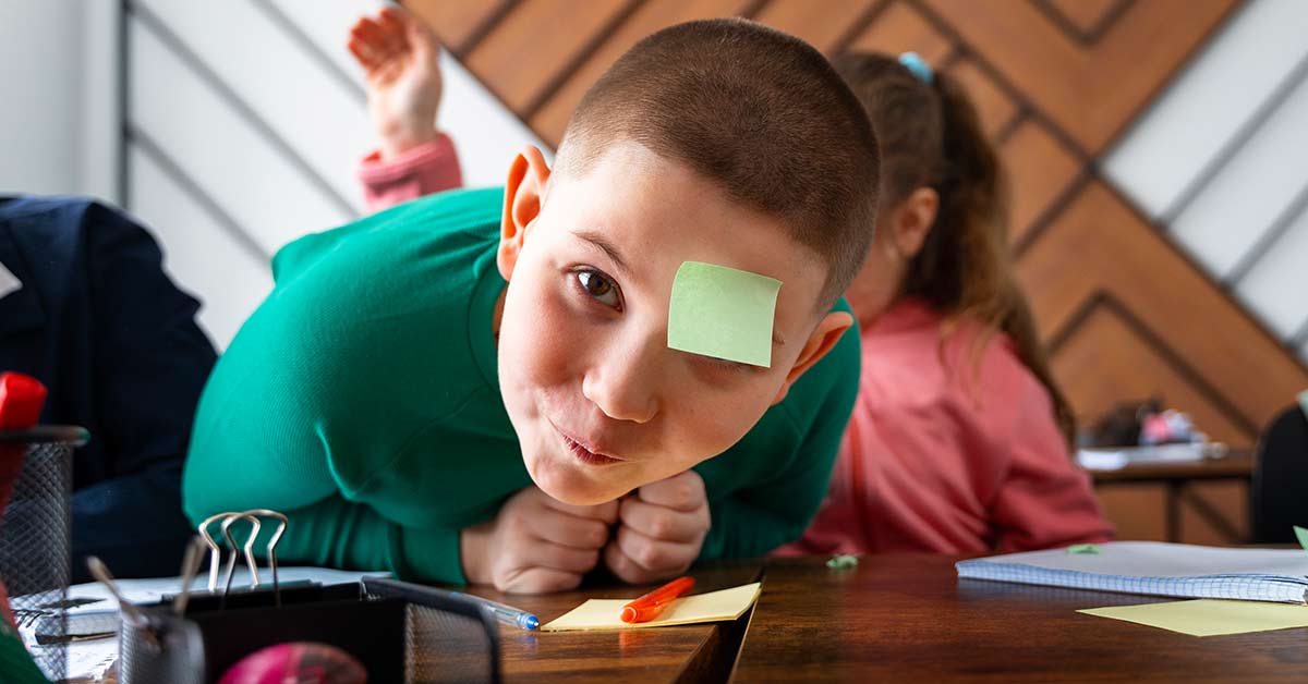 Criança sentada à secretária na sala de aula, com um post-it colado na testa e a mão levantada, num momento de brincadeira distraída